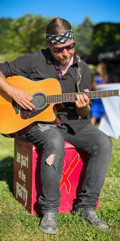 Gitarrist Toni sitzt auf einem roten Sitzwürfel auf einer Wiese und spielt Akustikgitarre, er trägt eine Jeans mit Rissen und hat um den Kopf ein Bandana gebunden, darunter einen Pferdeschwanz