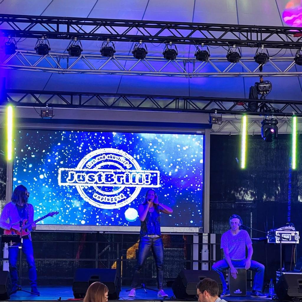 Die drei Musiker von JustBrill!-unplugged auf einer Open Air Bühne, dahinter groß das Logo der Band